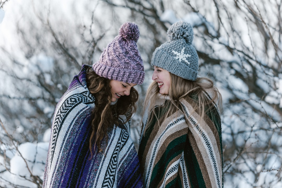 Two women warmly dressed in knit hats and colorful blankets stand close together, smiling and laughing in a snowy outdoor setting with bare trees in the background.