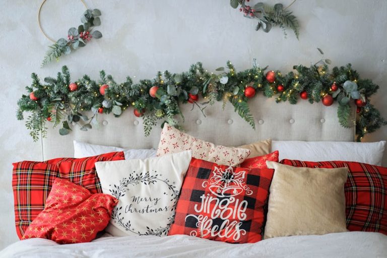 A cozy bed adorned with festive pillows featuring Christmas designs. Above the headboard, there's a garland decorated with red and gold ornaments, greenery, and fairy lights, creating a warm holiday atmosphere.