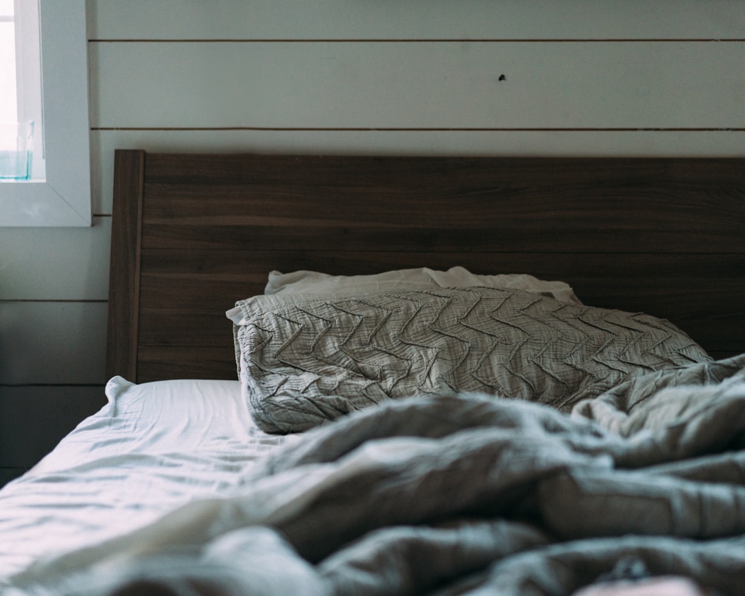 A cozy bed with a wooden headboard and patterned pillows is seen in a softly lit room. The bedding appears tousled, and soft light filters in through a window to the left, creating a serene and inviting atmosphere.