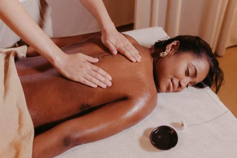A person is lying face down on a massage table, receiving a relaxing back massage. The setting is a tranquil spa environment, with a small dish of massage oil nearby. The person has a serene expression, and a towel covers their lower body.