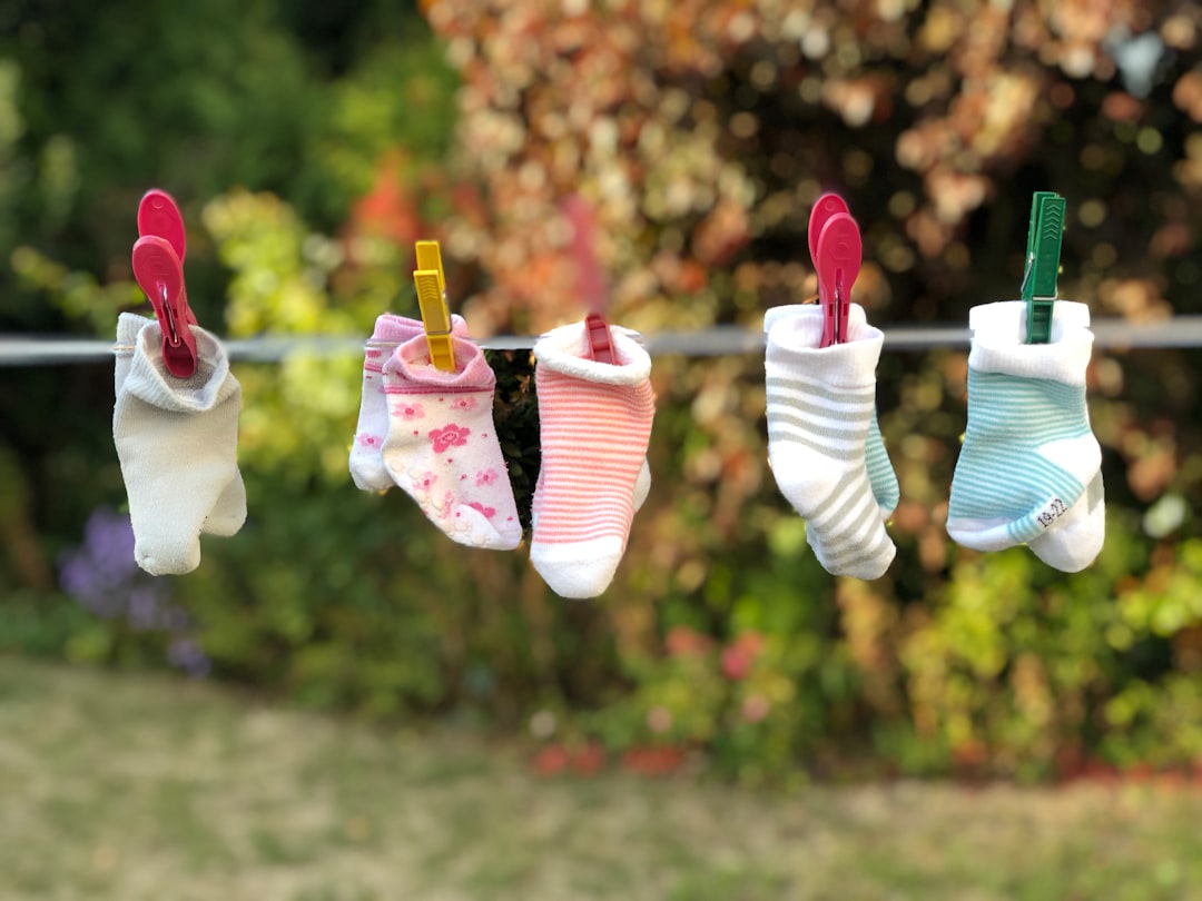 Five pairs of baby socks, each with different patterns and colors, are clipped with colorful clothespins to a clothesline outdoors, with a blurred background of greenery and flowers.