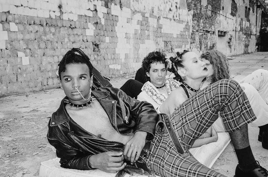 A group of people with punk fashion, wearing leather and plaid, sit and lie on the ground against a textured brick wall. Their styles include piercings, black lipstick, and unique hairstyles, exuding a rebellious, edgy vibe.