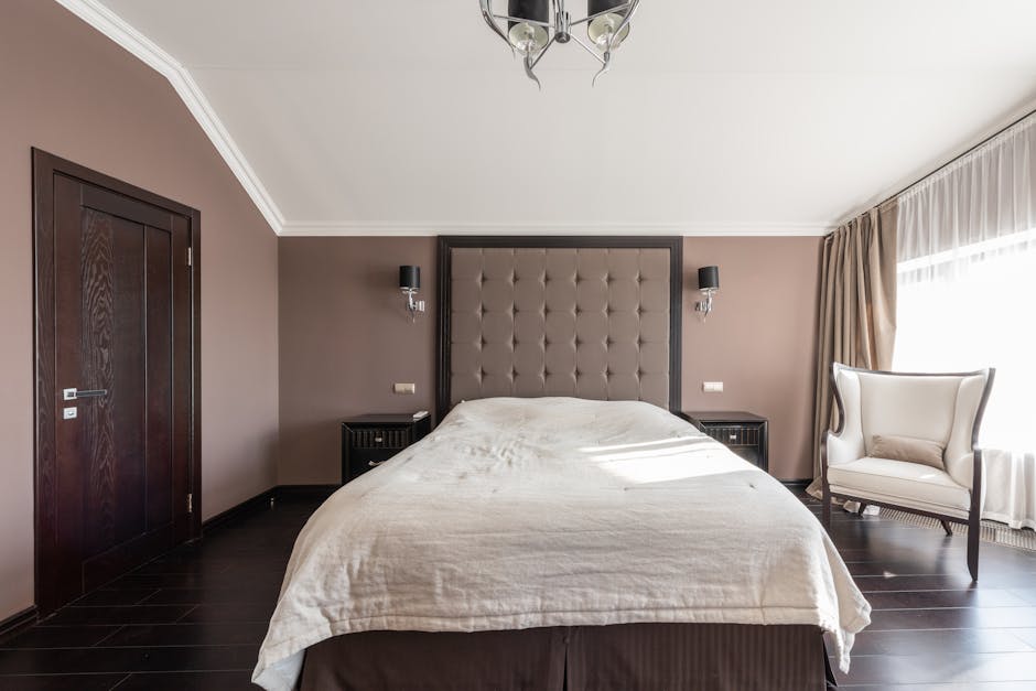 A modern bedroom with taupe walls and dark wood flooring. It features a large bed with a tufted headboard, two bedside tables with lamps, a white armchair by the window, and a black door. Natural light shines through the window with sheer curtains.