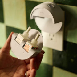 A hand holds a gray Pura device with two scent refills, while another is plugged in.