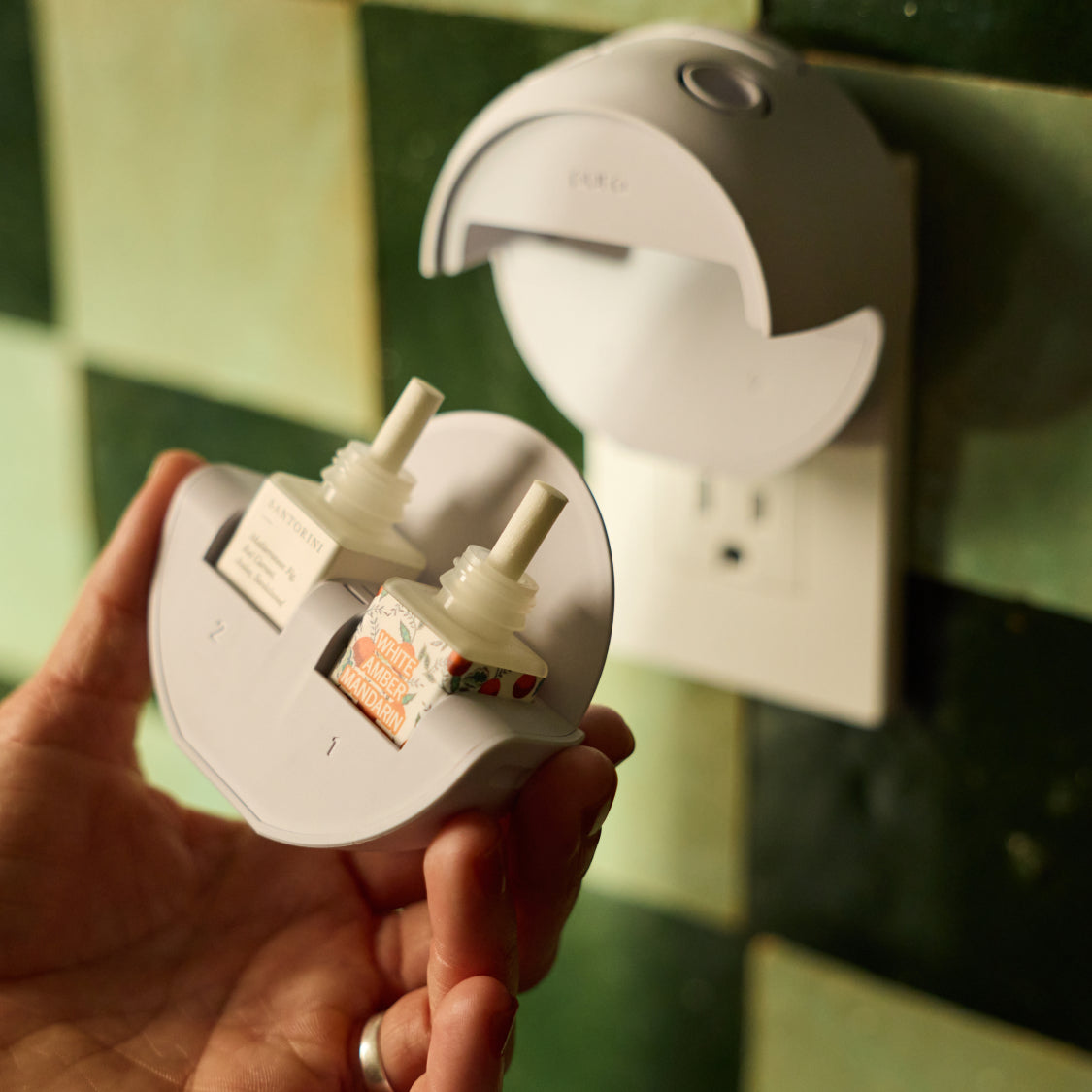 A hand holds a gray Pura device with two scent refills, while another is plugged in.