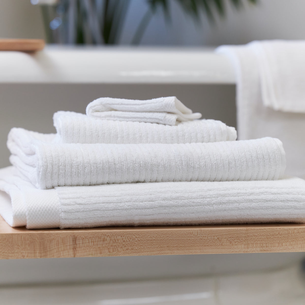 White Avocado organic cotton bath towels, hand towel, and washcloth stacked beside a bathtub